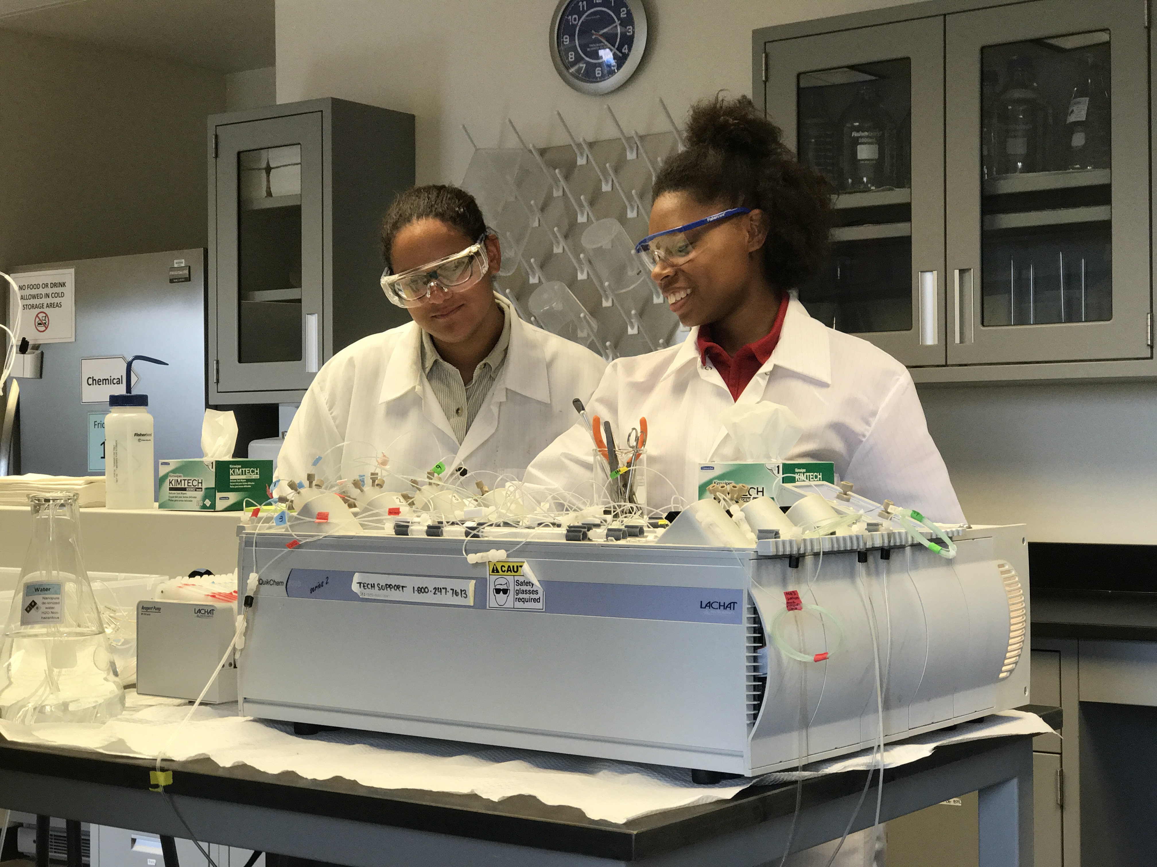 Two smiling college-age women in lab coats and protective eyewear conducting research in a laboratory. 