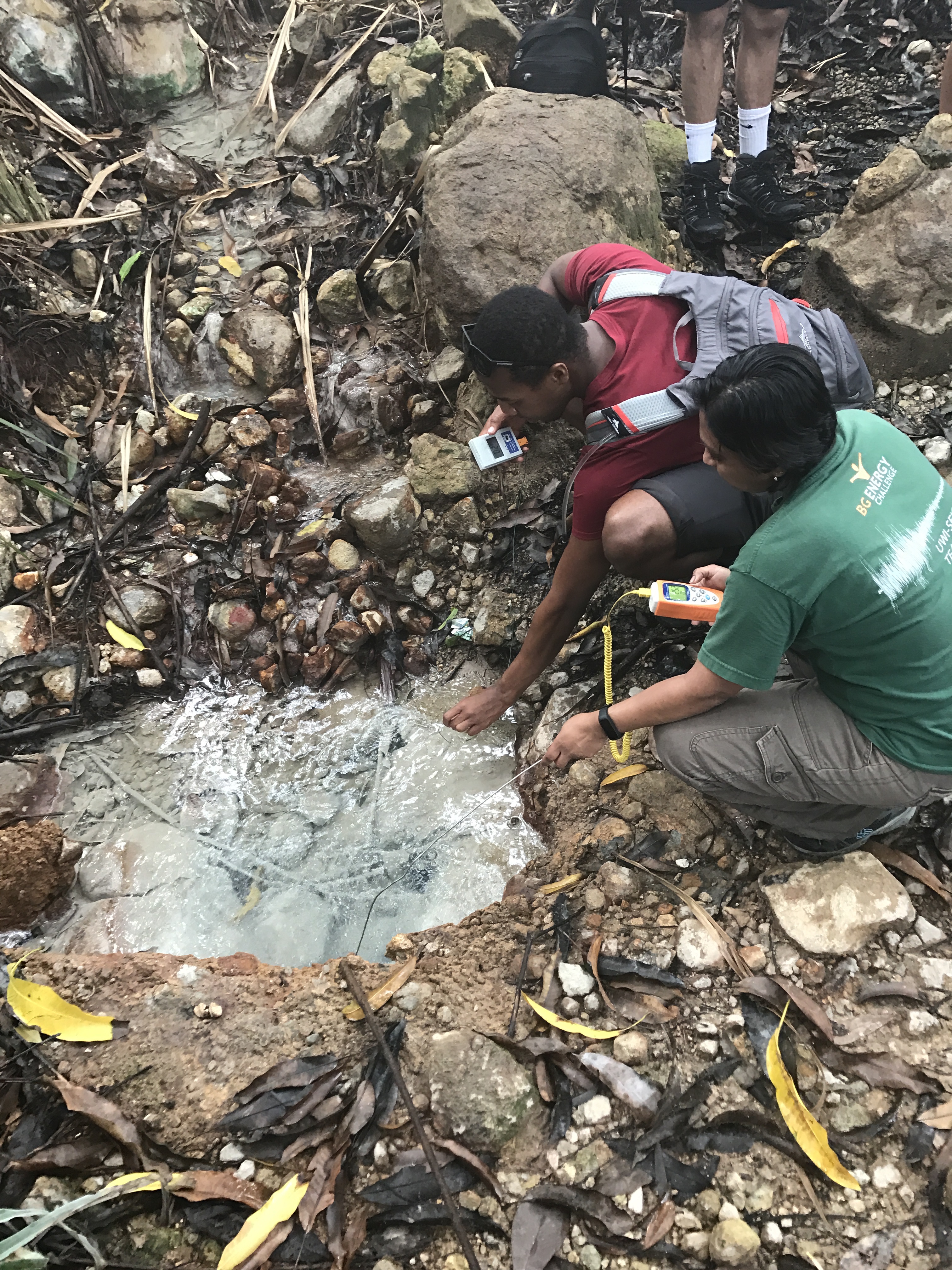 Two scientists crouch amongst boulders and leaves to collect water samples from a sinkhole.