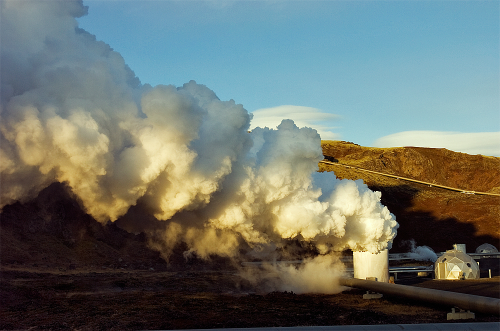 A geothermal power plant in a rocky, mountainous setting with steam billowing from one of the towers.