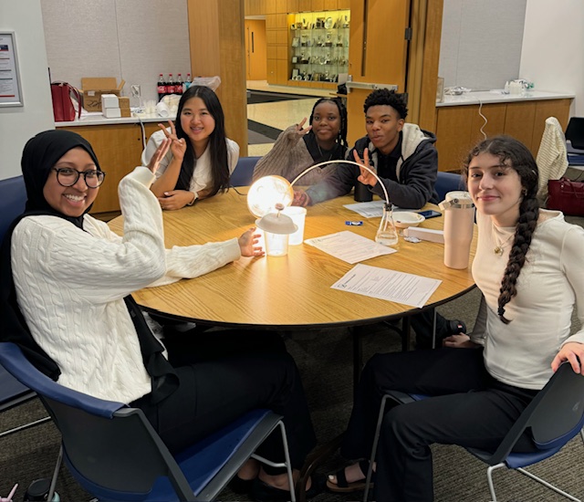 Five students pose around an energy-related project they have set up using a lamp and various containers.