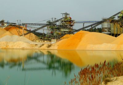 A body of water sits in front of piles of tan and grey sediment amongst mining equipment