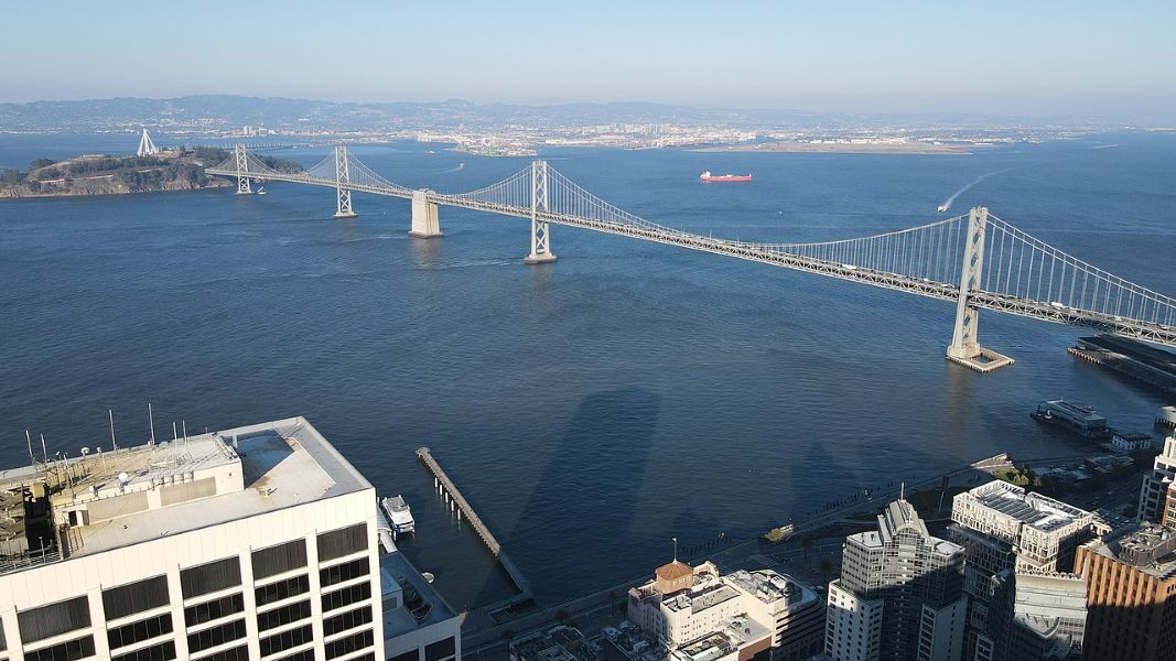 An aerial view of the San Francisco-Oakland Bay Bridge stretching across the bay, with calm blue waters and distant hills in the background. The image includes parts of downtown San Francisco's buildings in the foreground and a red cargo ship in the distance.