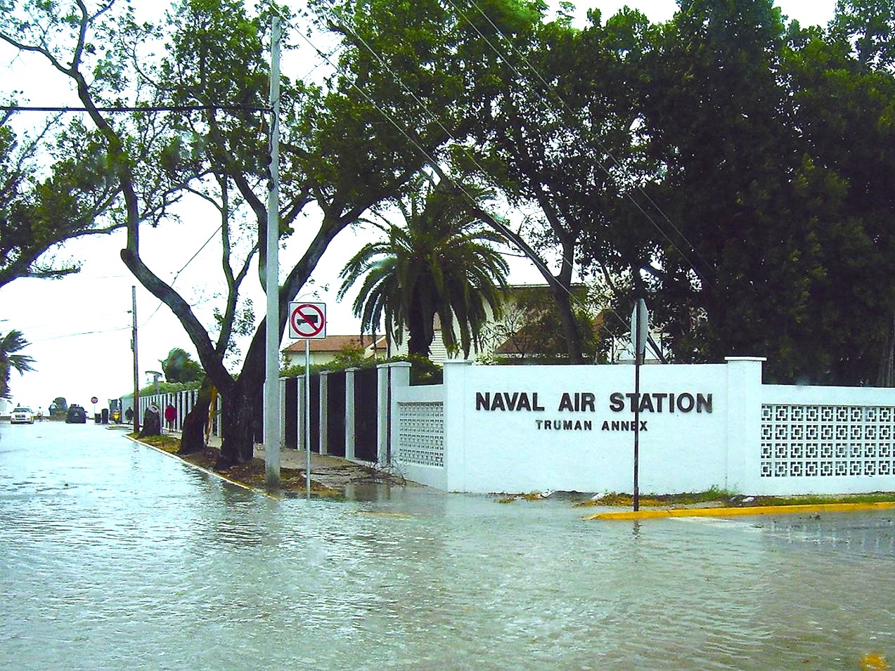 A flooded street outside the Naval Air Station Truman Annex, with standing water covering the roadway. Trees, a 'no left turn' sign, and a white perimeter wall with the station's name are visible, while cars and pedestrians navigate the waterlogged area.