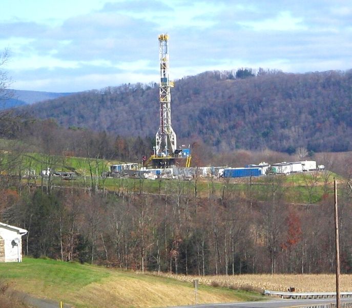 An image of a natural gas drilling rig set on a hillside with forested mountains in the background. The site includes industrial equipment, trucks, and storage containers, surrounded by leafless trees and grassy terrain under a cloudy sky.