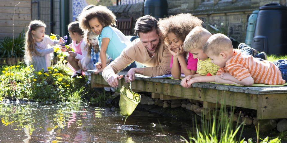 An adult with children on a boardwalk over a pond examine the contents of a scoop net.