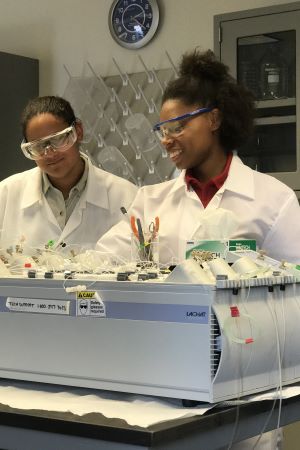 Two smiling college-age women in lab coats and protective eyewear conducting research in a laboratory.