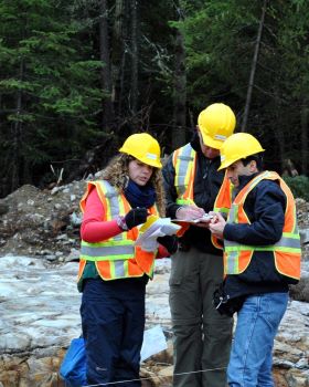 A woman and two men, all in outdoor safety vests and hardhats, stand on a rocky area discussing notes.